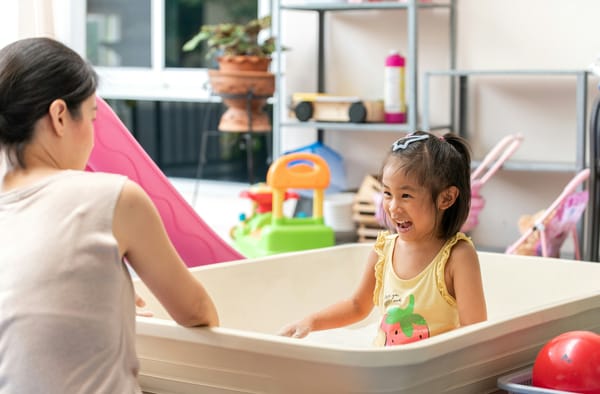 A girl with dark hair and a yellow tank top plays in a large white indoor sandbox with an adult nearby.