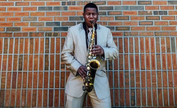 Black man in tan suit playing saxophone with eyes closed in front of brick building