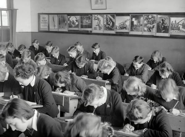 Black and white photo of white children in school uniforms bent over written coursework